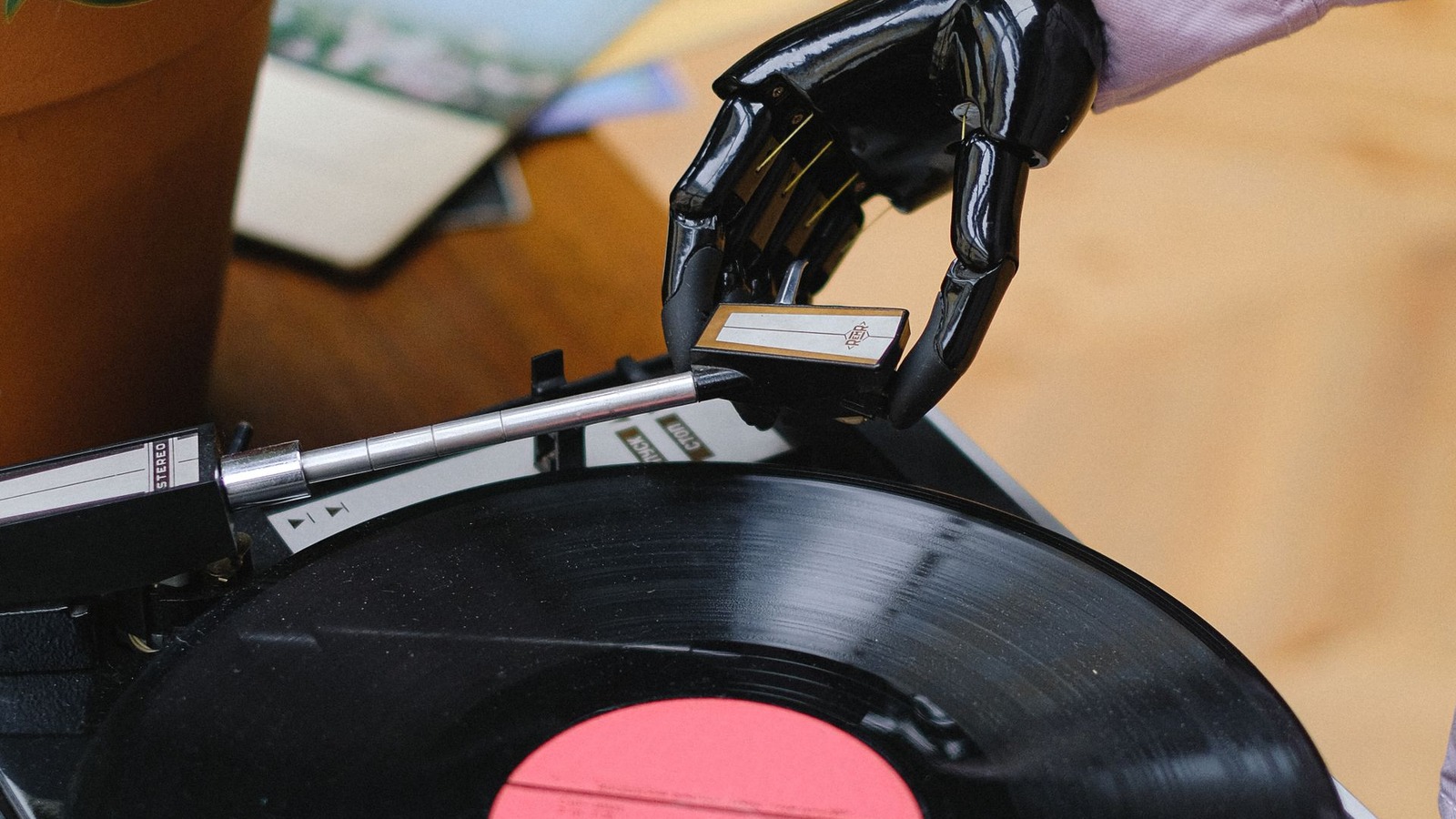 Mana robotica neagra aseaza acul unui pick-up pe un disc de vinil cu eticheta rosie — muzica generata cu inteligenta artificiala