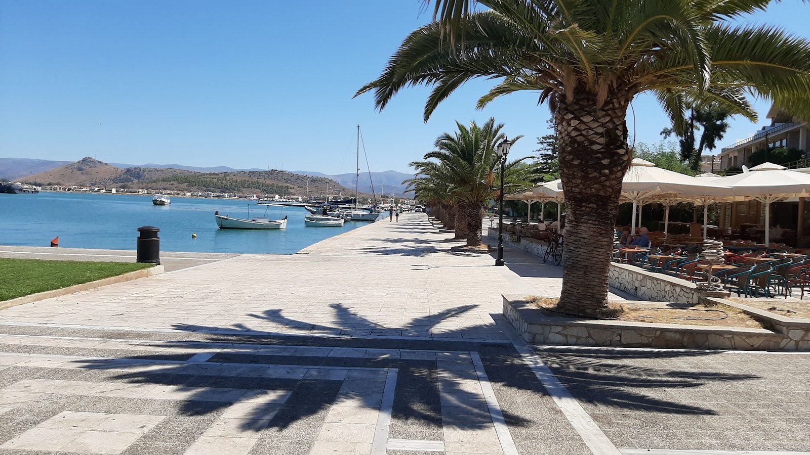 Faleza unui port grecesc: palmieri cu umbre lungi pe pavaj, bărci de pescari pe apă turcoaz, taverne cu umbrele albe în dreapta și dealuri în fundal.